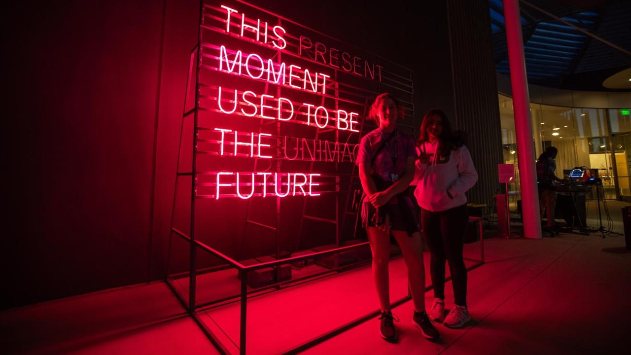 Two people standing in front of neon text artwork by Alicia Eggert with the phrase "This moment used to be the future" illuminated.