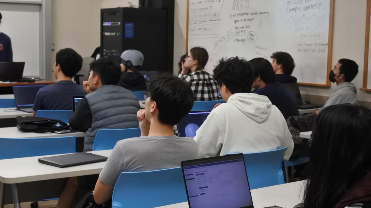 the backs of several students seated in a classroom looking toward the front of the room, where a presentation is being held.