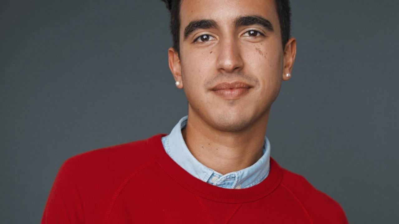 A man with dark, coifed hair wearing a red sweater stares directly at the camera