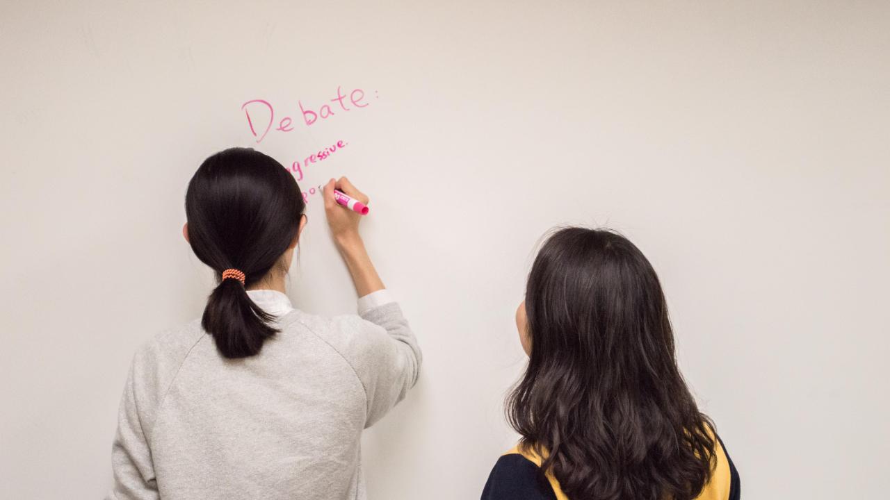 Two students write debate and other words on a whiteboard.