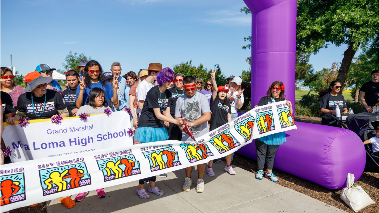 A crowd of people is gathered behind an inflatable purple archway. Two people in the center of the photo are cutting a ribbon patterned with the Best Buddies logo. They are beginning the walk course.