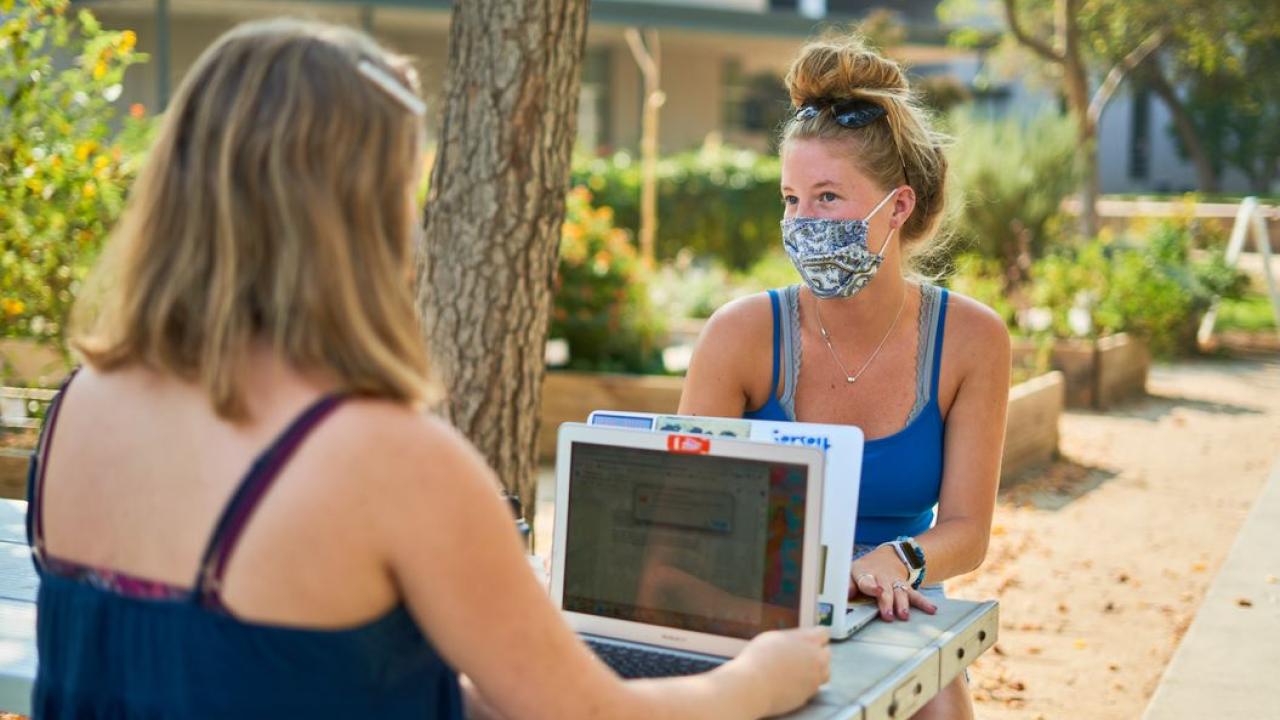 Two students at outdoor table working on laptops with face masks on