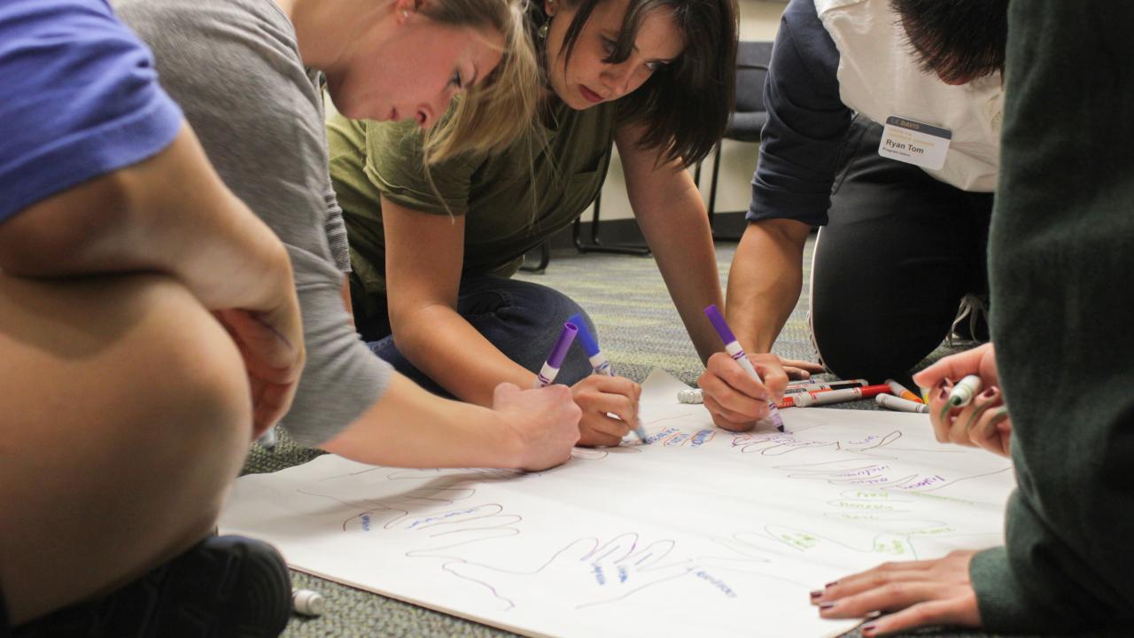 group of students writing with markers on large sheet of paper on the floor