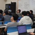 the backs of several students seated in a classroom looking toward the front of the room, where a presentation is being held.