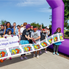  A crowd of people is gathered behind an inflatable purple archway. Two people in the center of the photo are cutting a ribbon patterned with the Best Buddies logo. They are beginning the walk course.